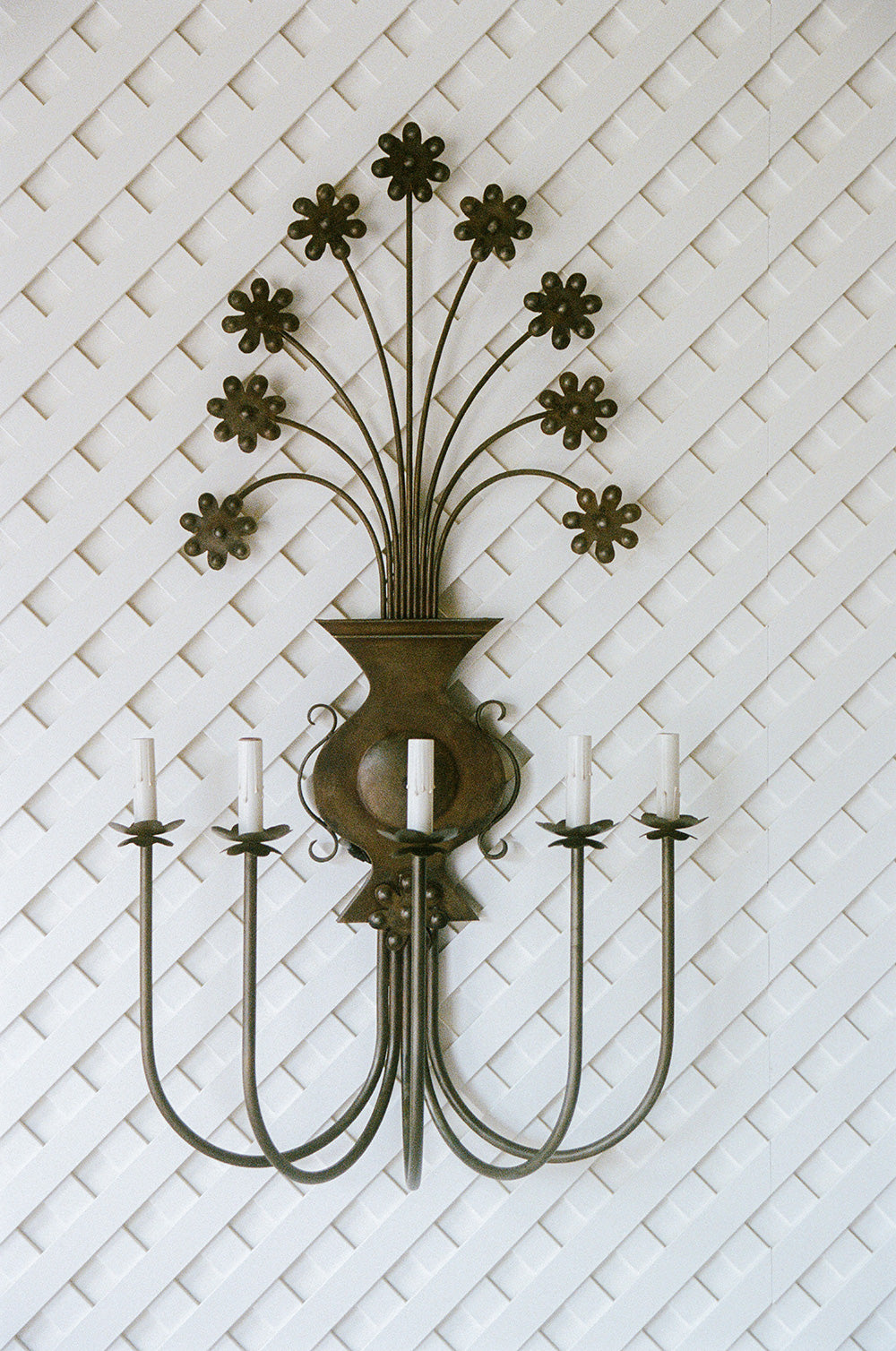 Bronze patina iron wall sconce with five candle arms below a spray of flat daisy flowers rising from an urn, against white lattice