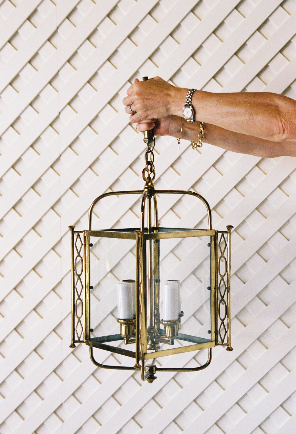 Brass four light lantern with glass panels, held up by its chain against a white lattice wall