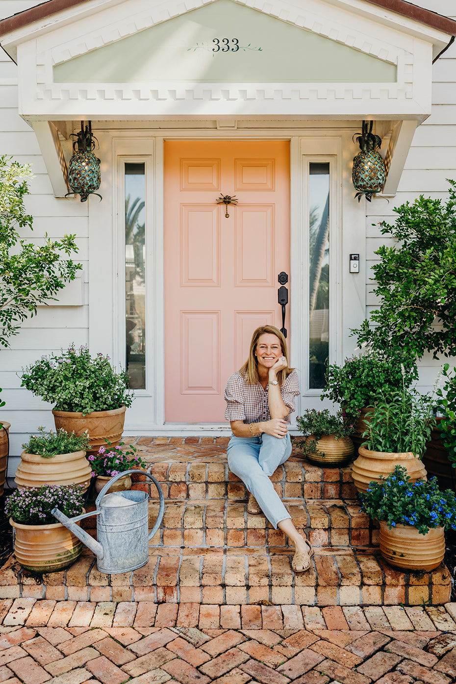 Cece Bowman sitting on brick steps in front of a pink front door, surrounded by potted plants