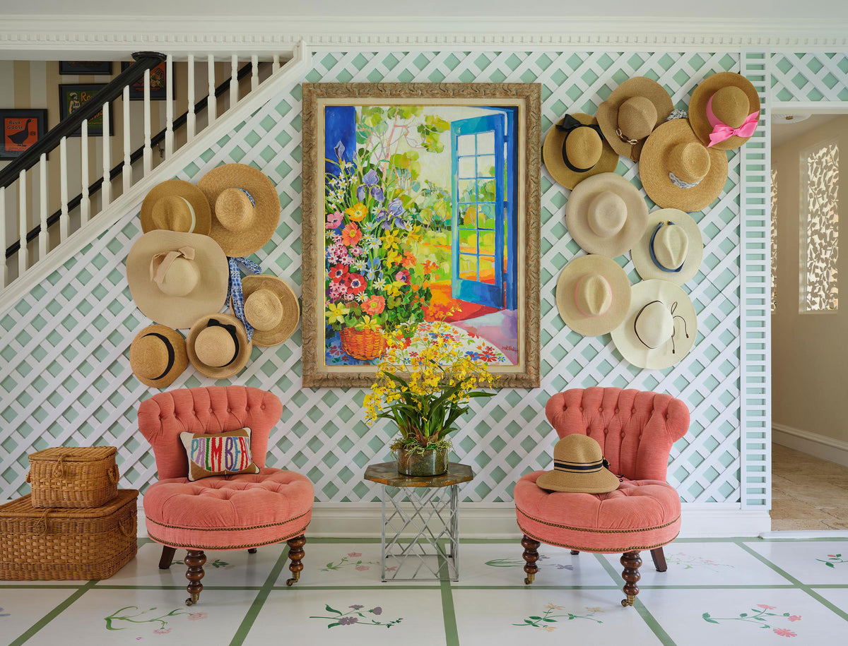 Entry hall with straw hats hung on green trellis wallpaper, a floral painting and two coral tufted chairs