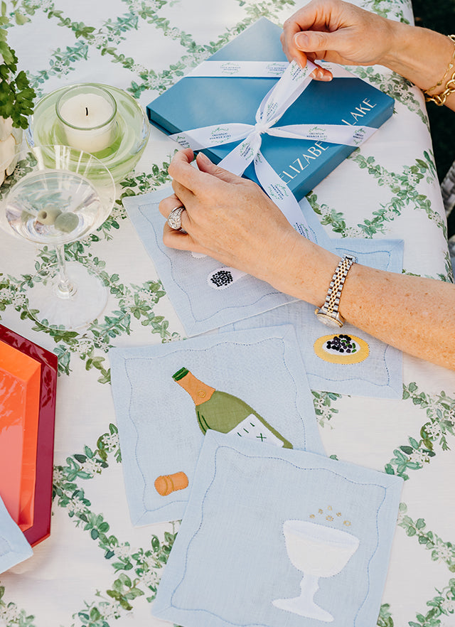 Hands tying a blue Elizabeth Lake gift box over embroidered cocktail napkins on a green trellis tablecloth