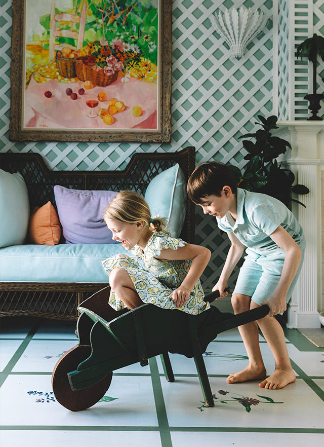 Two children playing with a toy wheelbarrow in a sunroom with a green lattice wall