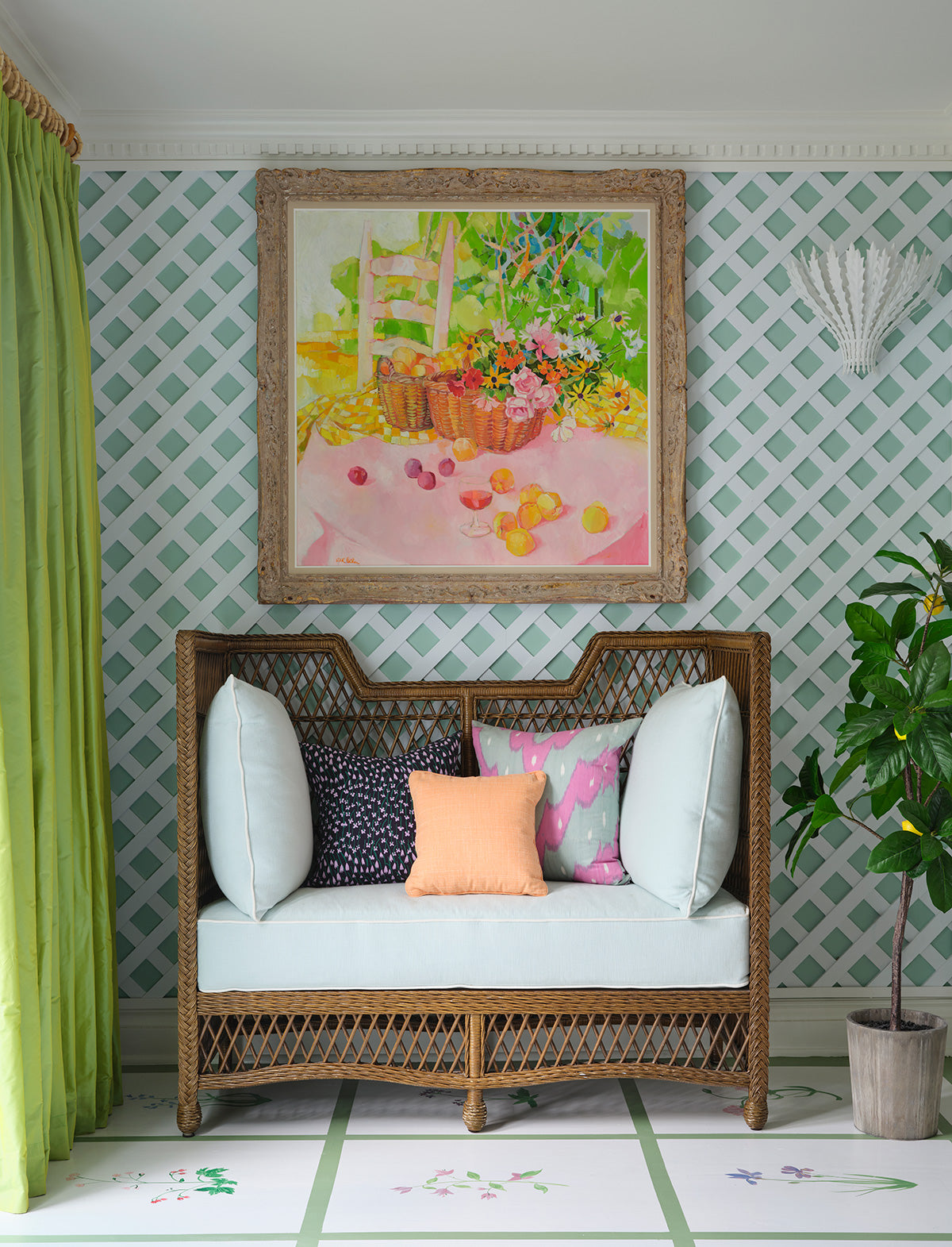 Wicker settee with pale blue and pink cushions beneath a still life painting, against green trellis wallpaper with a green curtain and a potted citrus tree