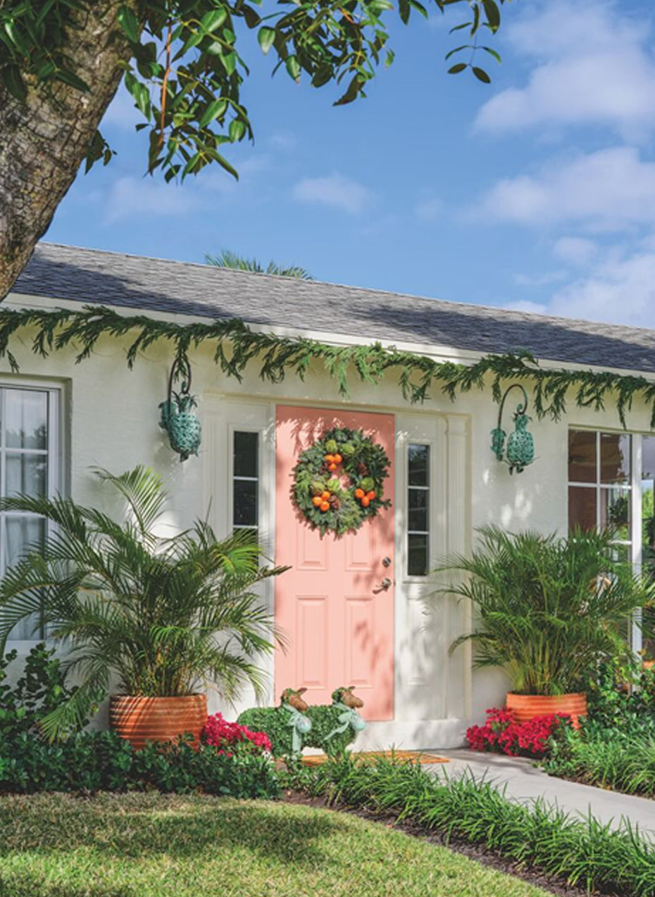 White cottage with a coral front door, a wreath and garland, and tropical plantings