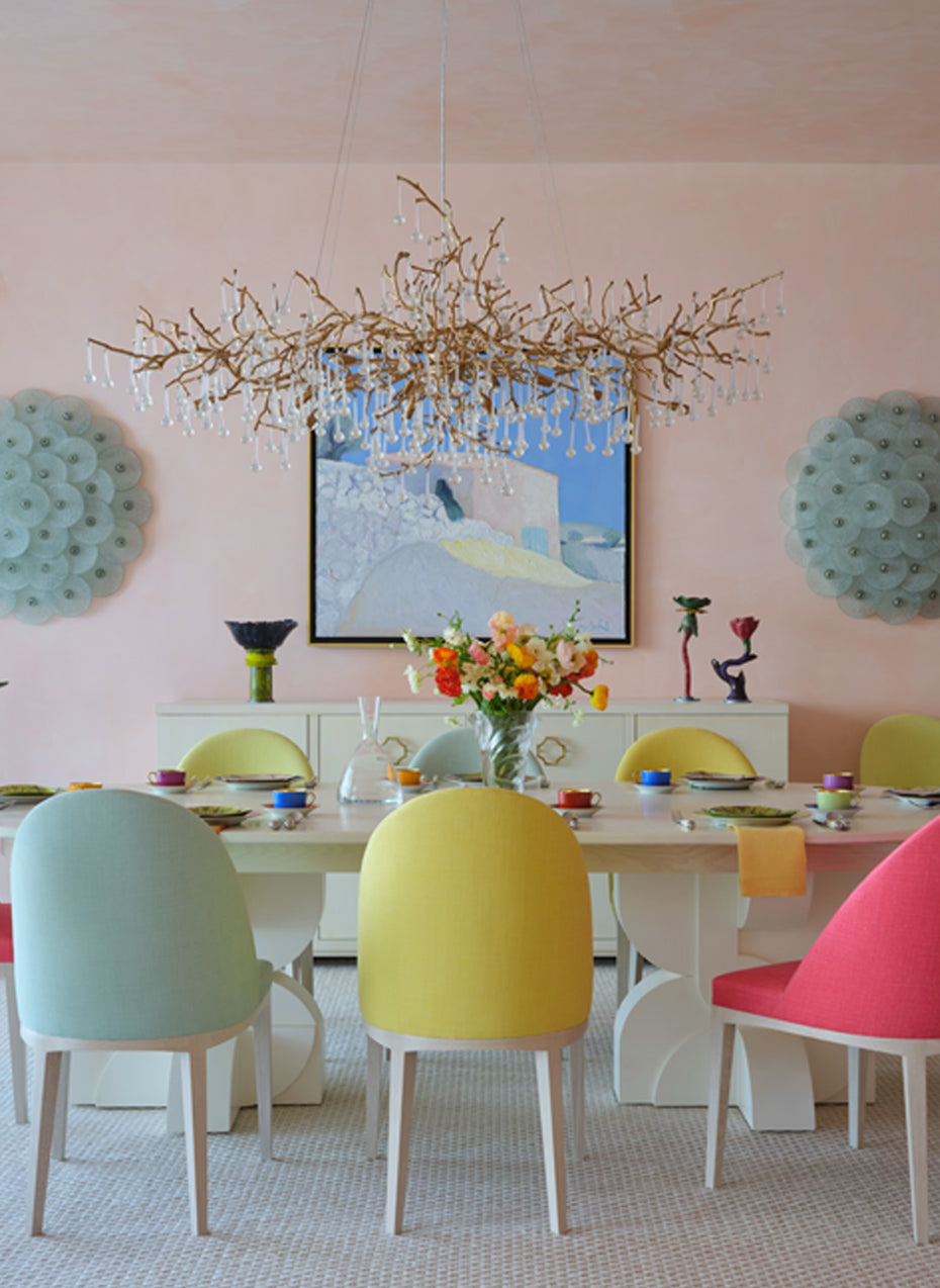 Blush pink dining room with pink, yellow and aqua chairs beneath a gilded branch chandelier