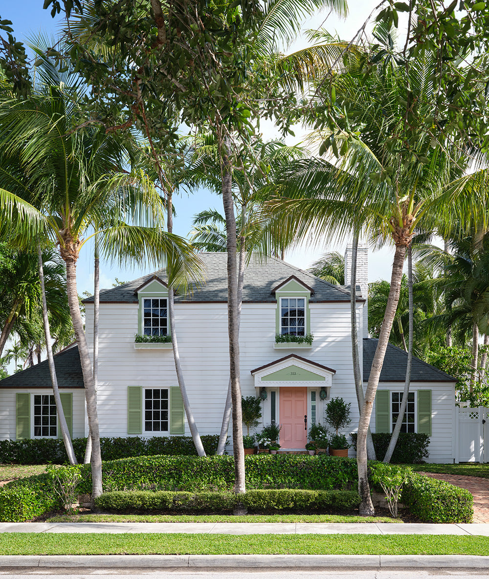 White two storey house with green shutters and a pink front door among palm trees
