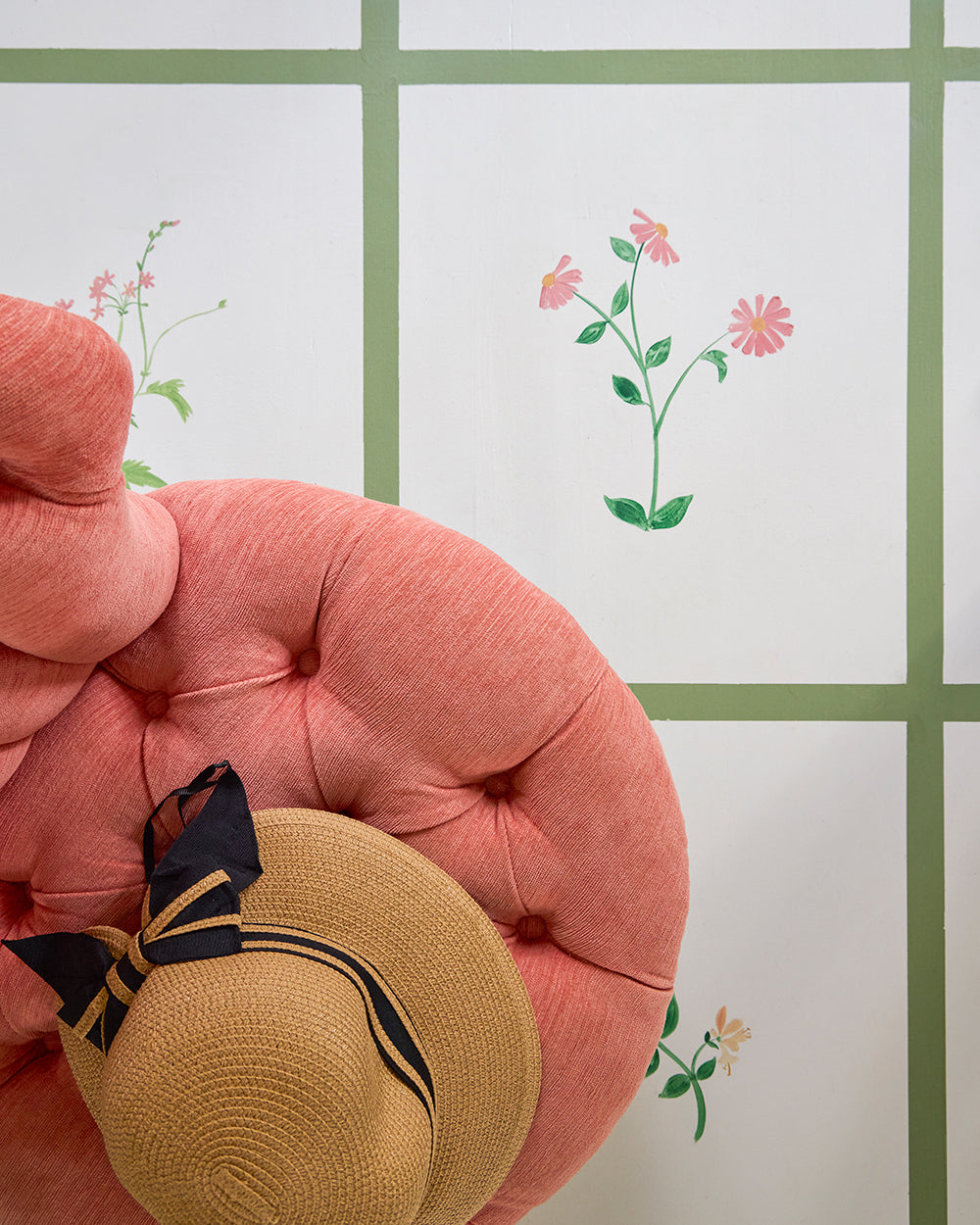 Coral tufted chair and a straw hat against a painted floral wall with green trim