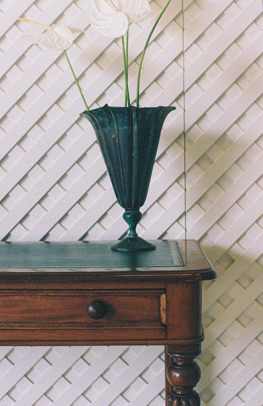 Fluted verdigris metal vase holding three white anthuriums, standing on a leather topped writing table