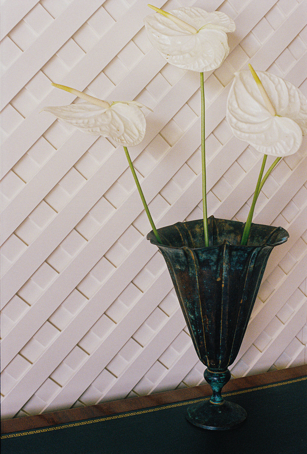 Fluted verdigris metal vase on a footed stem, holding three white anthuriums against white lattice