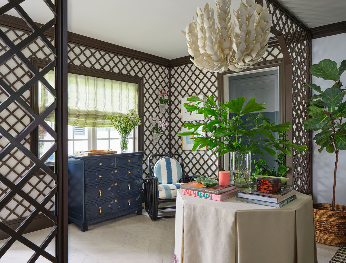 Sitting room framed by dark trellis panelling, with a paper lantern pendant, a navy chest and a skirted round table of books beside a fiddle leaf fig
