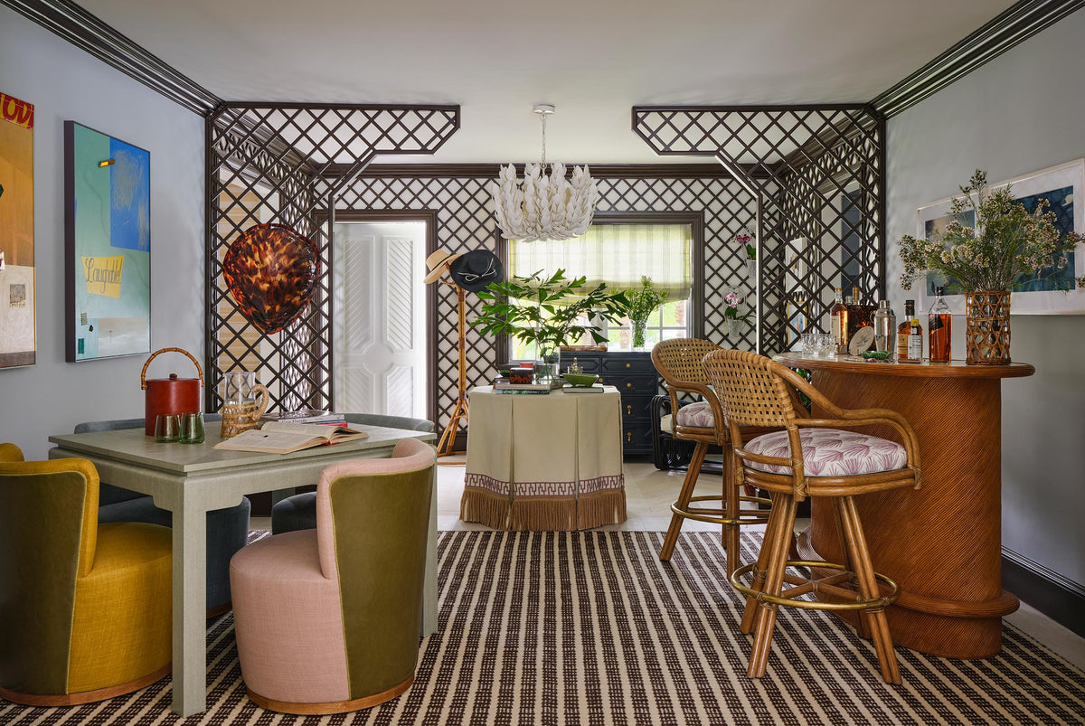 Games room seen through trellis walls, a grey card table with mustard and pink swivel chairs, a rattan bar with caned stools and bright canvases