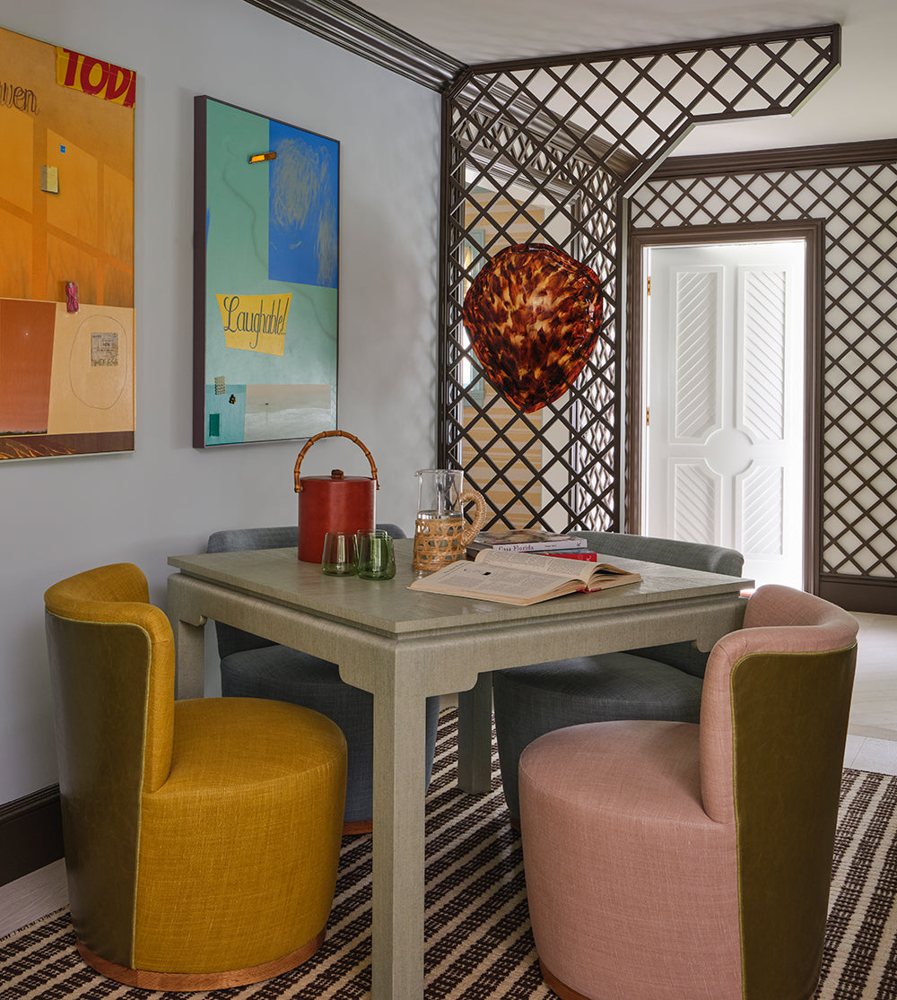 Grey card table with mustard and pink swivel chairs on a striped rug, bright canvases and a shell sconce on the trellis wall