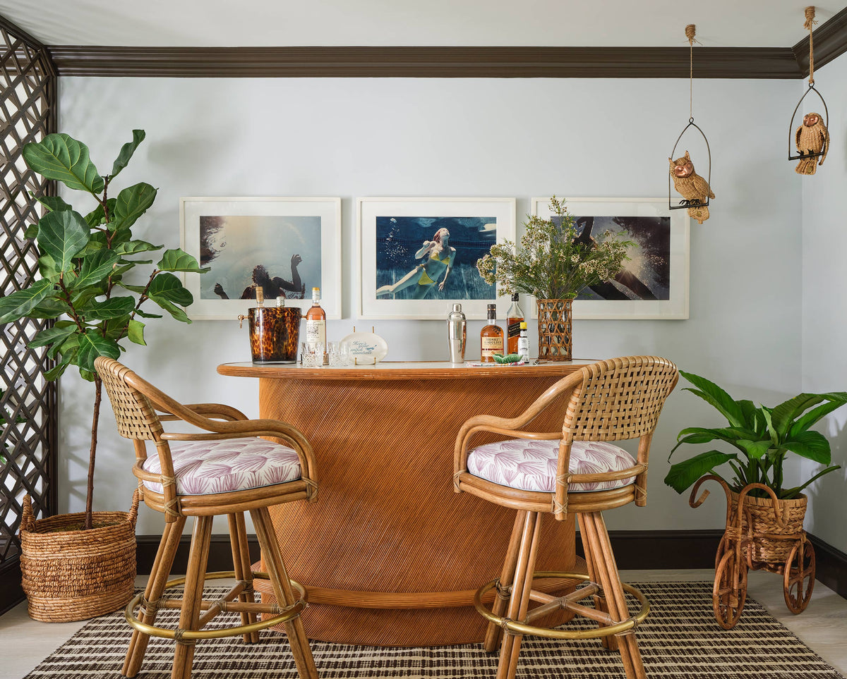 Curved rattan bar with caned stools under owl sconces, framed photographs and potted figs against a trellis wall