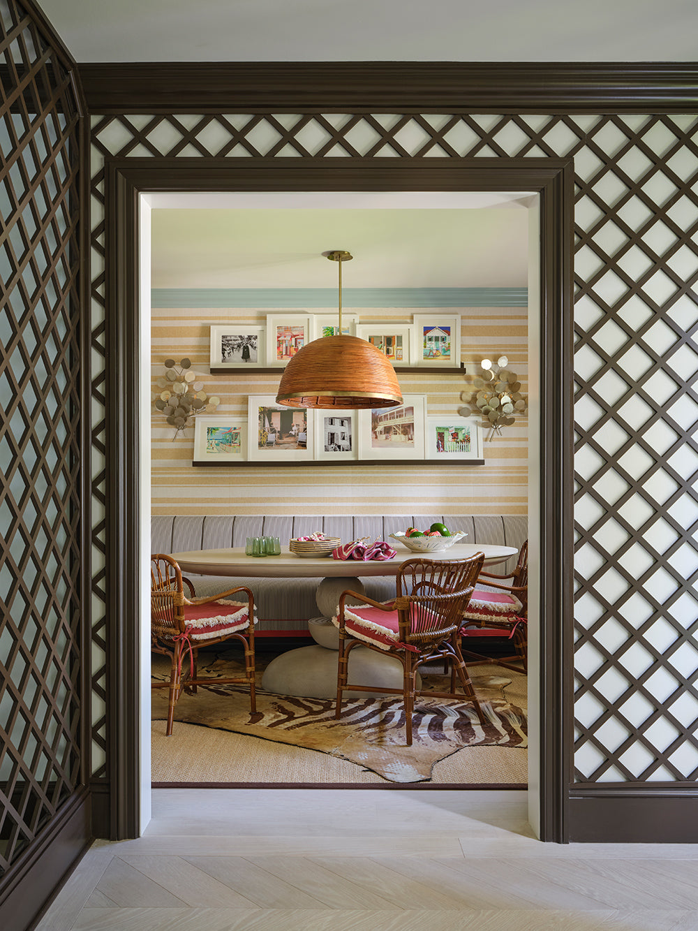 View through a dark trellis doorway to a breakfast room with a rattan dome pendant, a picture shelf and cane chairs on a zebra rug