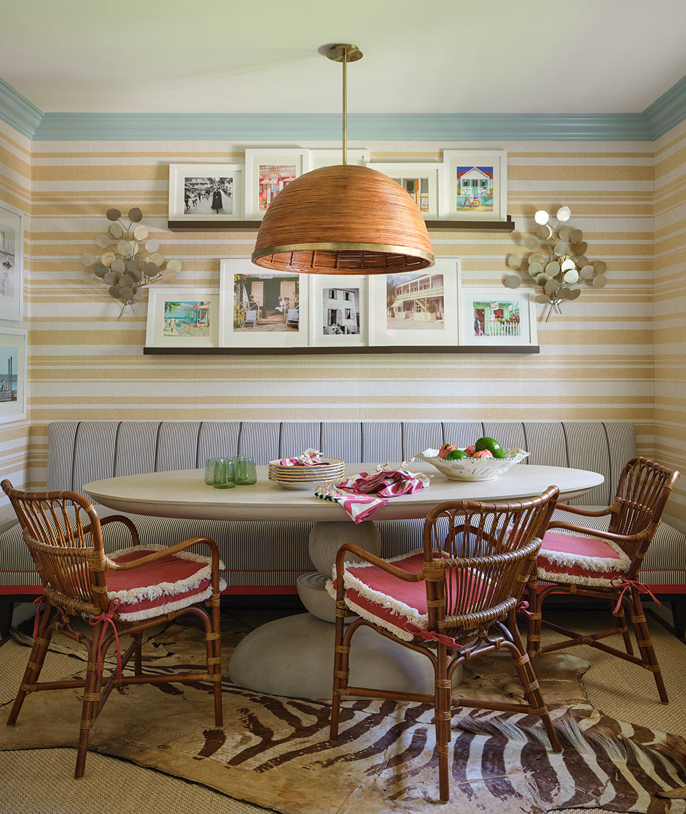 Breakfast room in striped grasscloth with a rattan dome pendant, framed art on shelves, a banquette and cane armchairs with pink cushions on a zebra rug