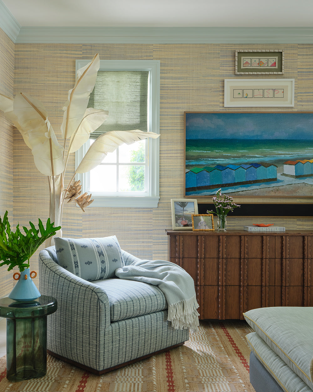 Corner with a blue and white striped swivel chair, a coastal painting over a rattan chest, a dried palm frond and a green glass side table