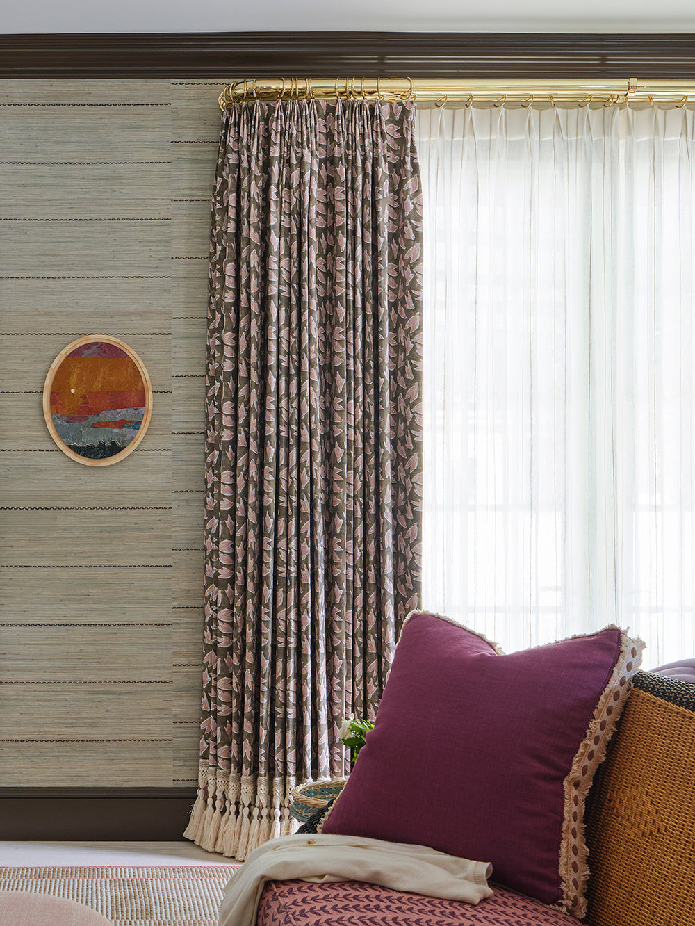 Curtain detail, brown patterned drapery on a brass rod over sheer panels, with a magenta cushion and a small oval painting