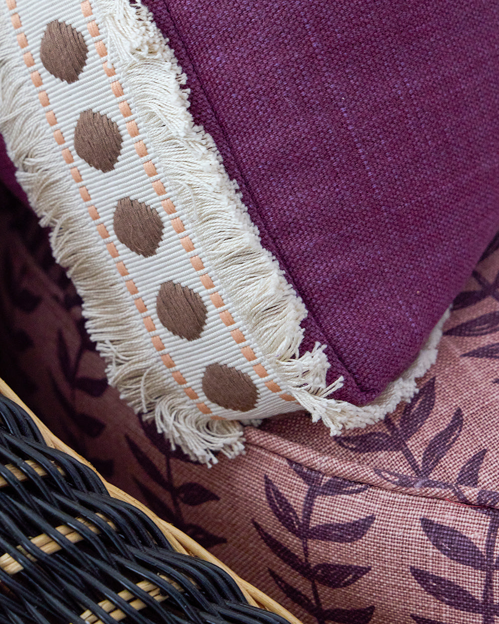 Close detail of a magenta linen cushion with fringed and dotted tape, over navy wicker and a purple leaf print