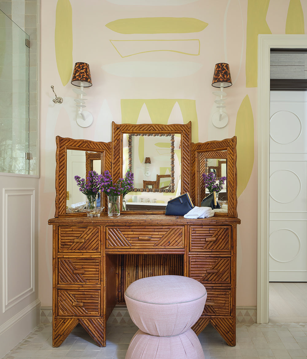 Rattan vanity table with a triptych mirror, scalloped lamps and a pink stool against yellow patterned walls