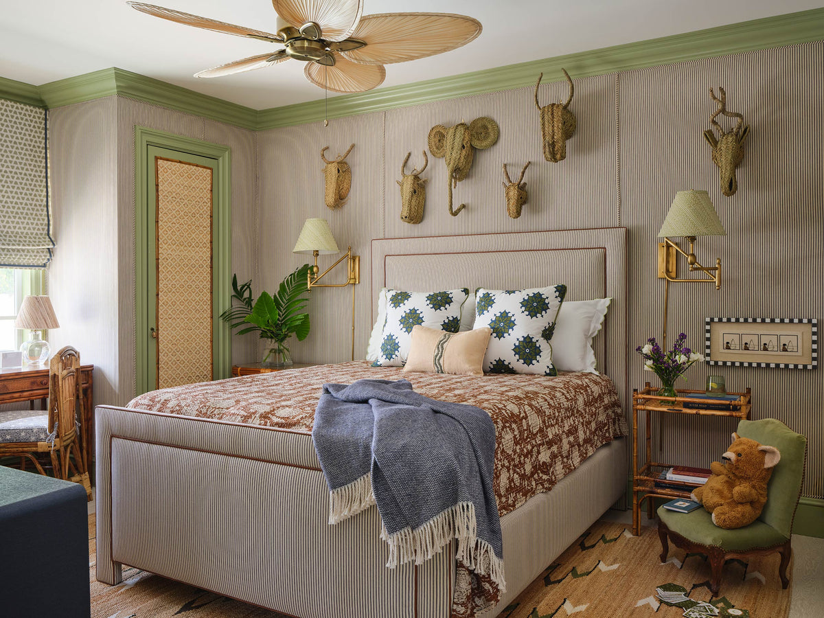 Bedroom with an upholstered bed under a brown floral coverlet, rattan sconces and a collection of woven animal heads on the wall