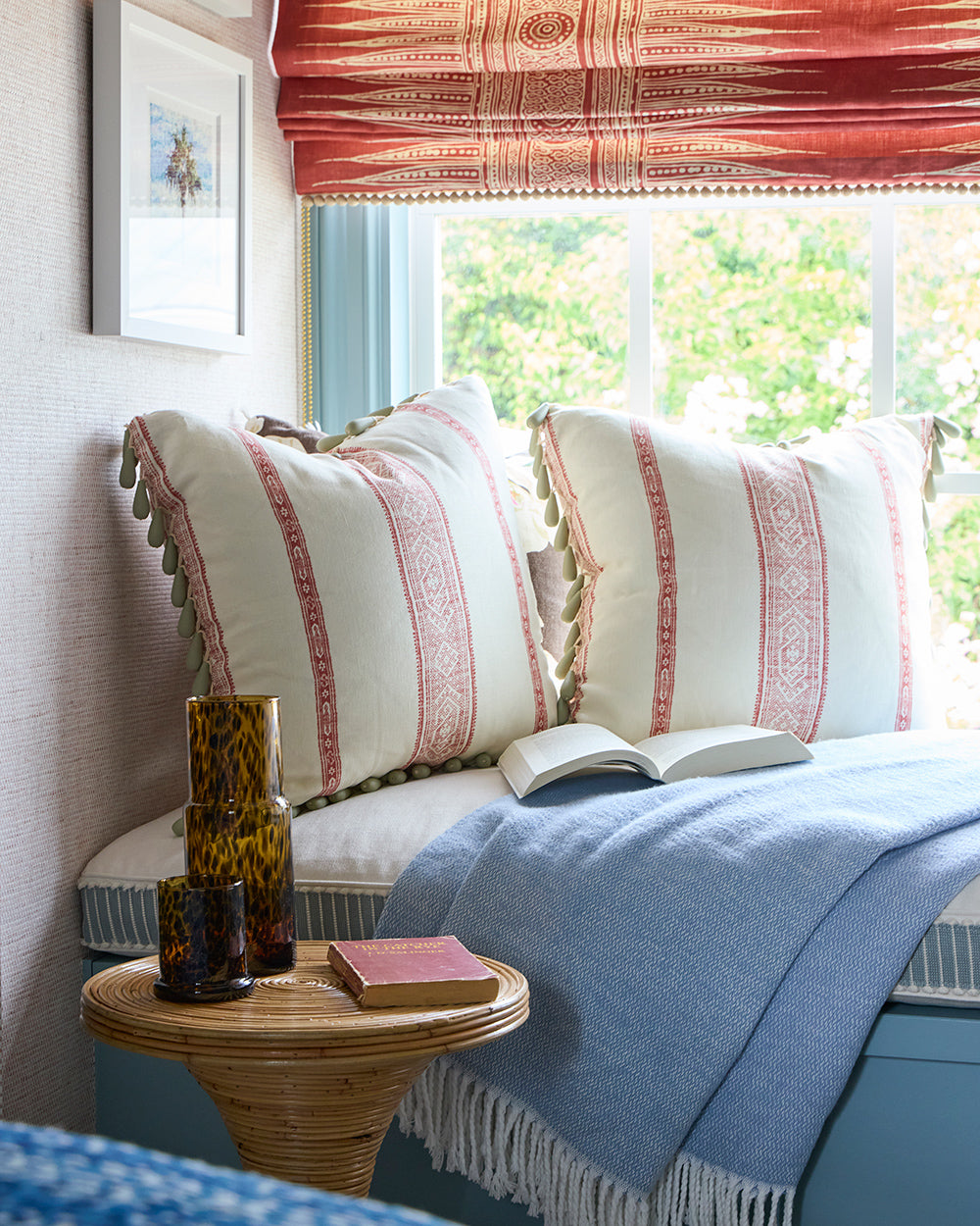 Window seat with striped and tasselled cushions, a blue throw and a rattan side table holding books