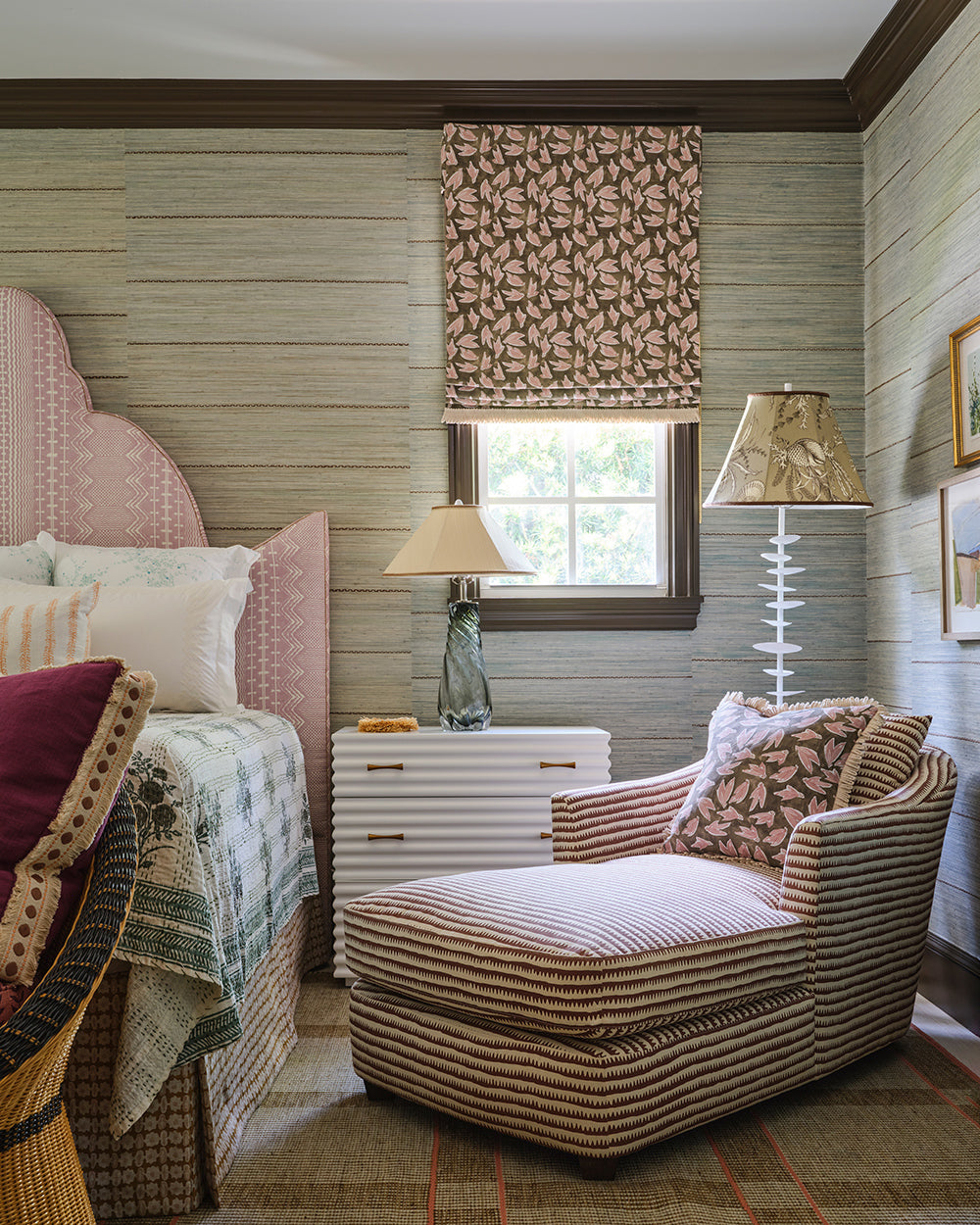 Bedroom corner in grasscloth with a pink scalloped headboard, a brown patterned roman shade and a striped chaise beside a woven side table