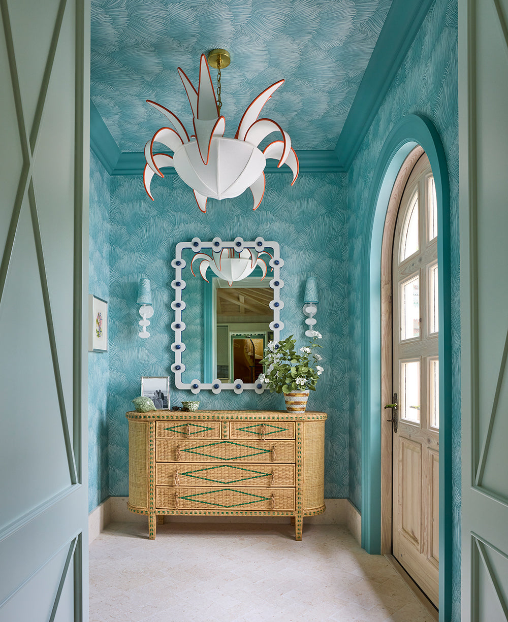 Entry hall in turquoise palm print wallpaper with a white fretwork mirror over a caned chest, an arched door to the garden