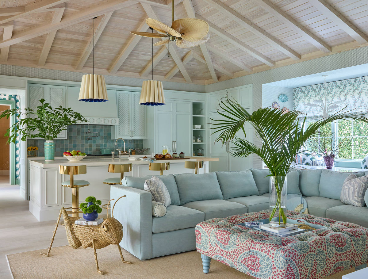 Open plan kitchen and sitting room under a pale timber vaulted ceiling, with pleated pendants, a grey sectional and a pink patterned ottoman