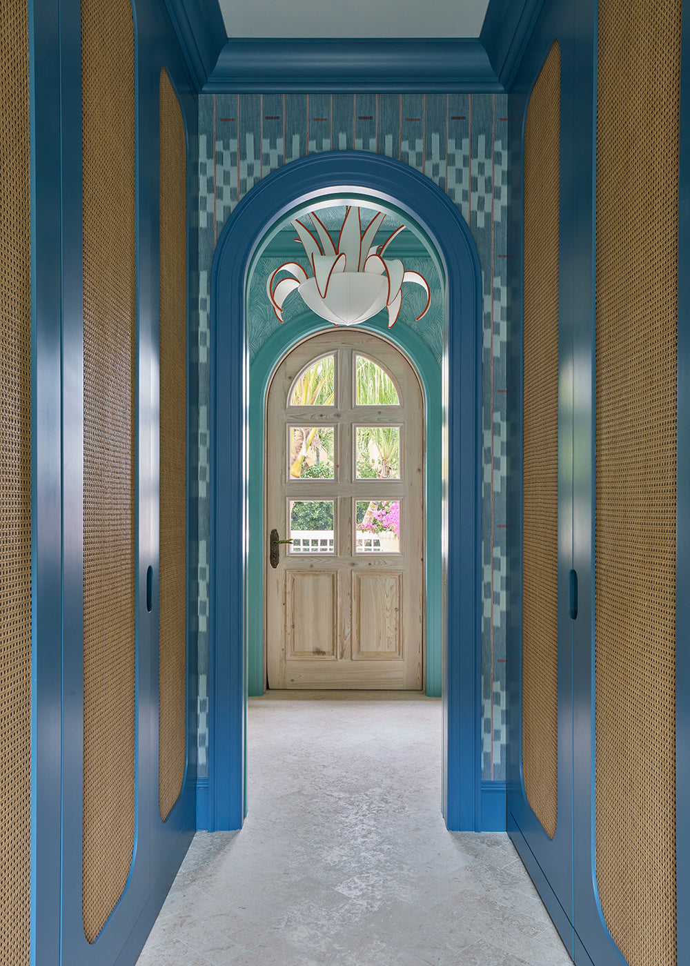 Corridor in blue grasscloth and caned panels leading to an arched front door under a white lily pendant