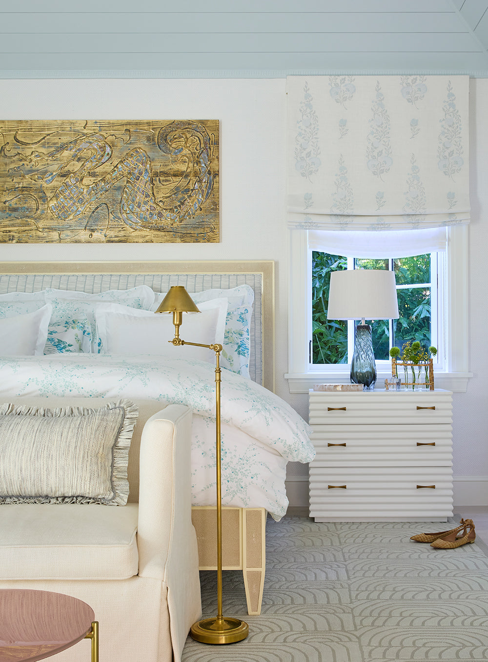Bedroom with a gilt relief above the bed, white bedding, a brass floor lamp and a printed roman shade