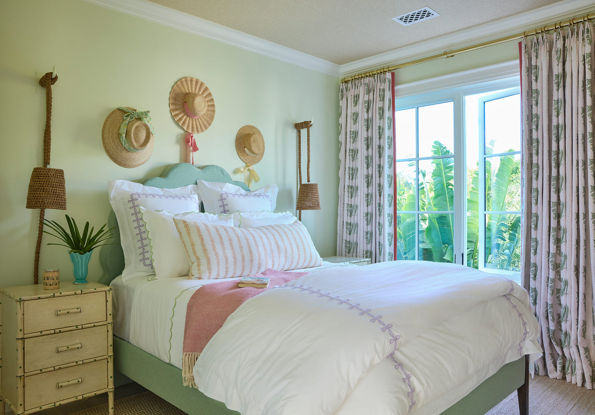 Bedroom in pale green with woven hats on the wall, a green bed frame, white bedding and printed curtains