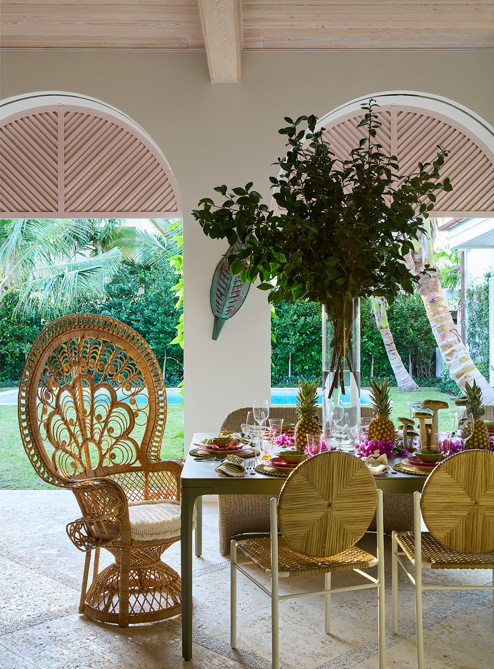 Loggia dining table under a topiary, with a peacock wicker chair and fan backed rattan chairs