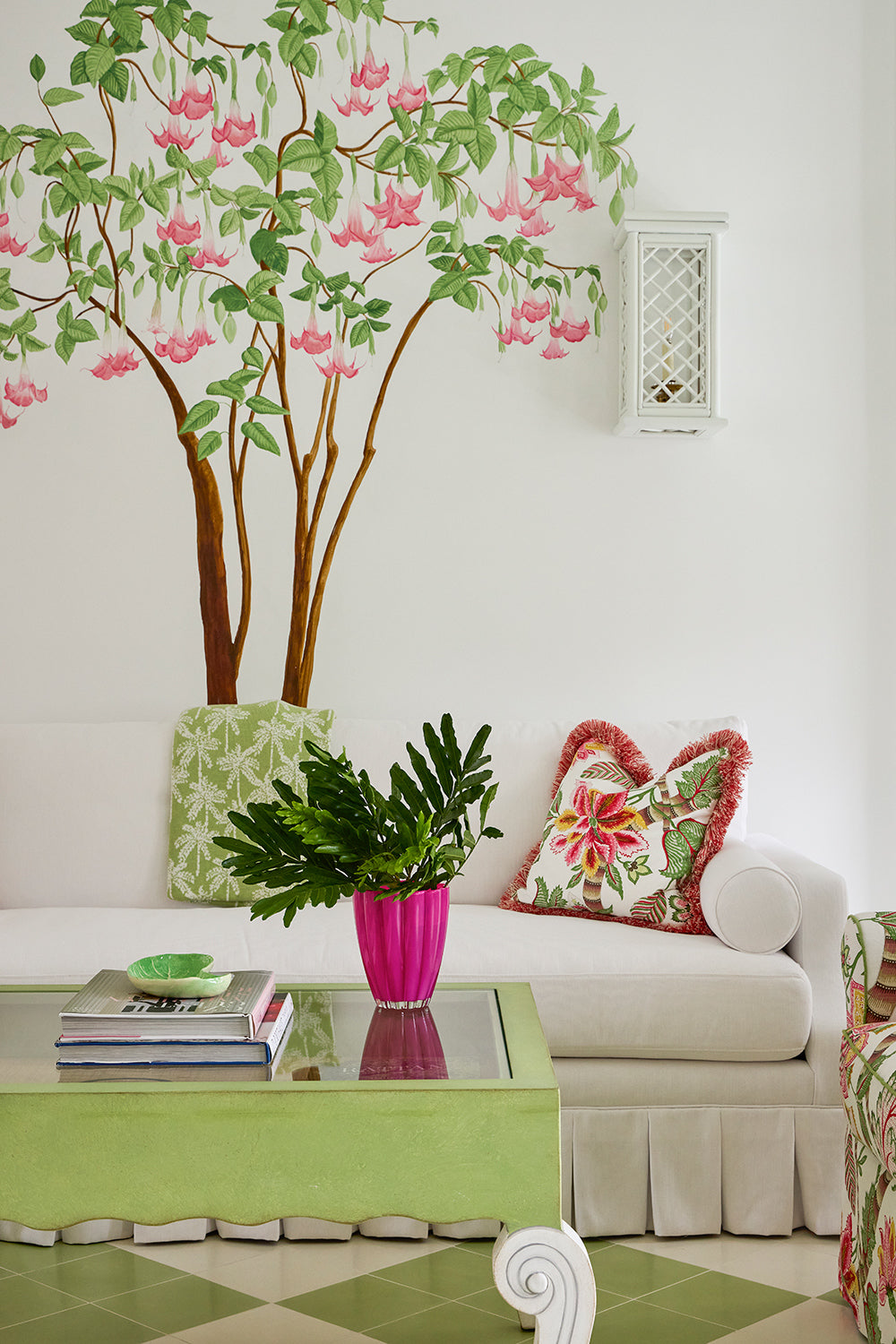 Corner with a hand painted flowering tree mural above a white sofa, a green skirted ottoman and a pink pot of leaves on a stack of books