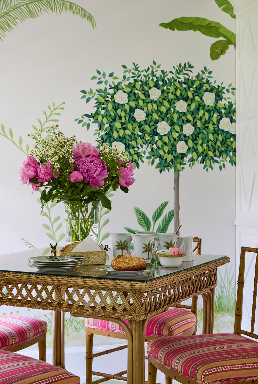 Rattan table laid with dishes under a painted topiary mural, pink peonies in a glass vase and striped bamboo chairs