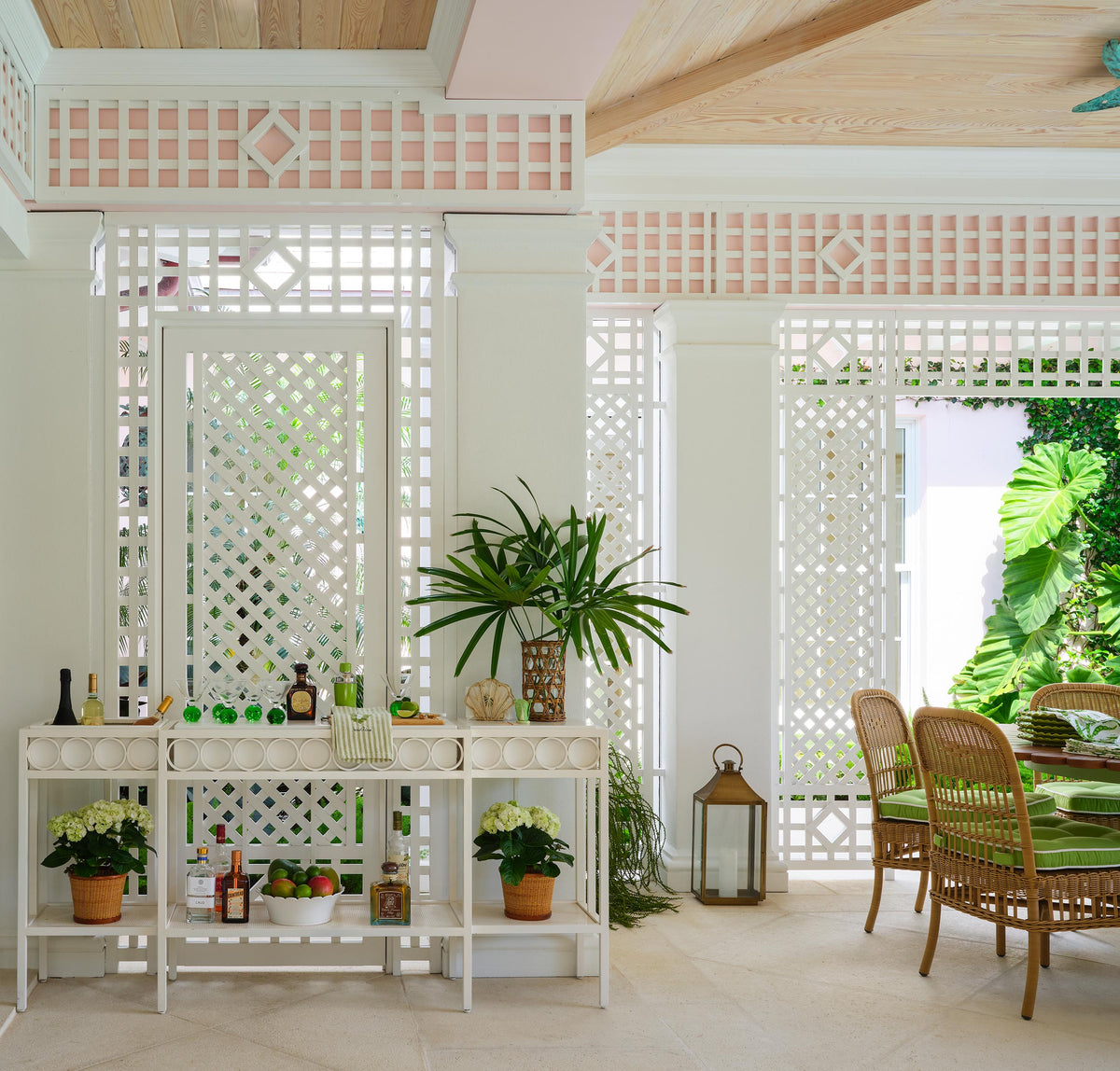 Lattice pavilion with a white console set as a bar, potted palms and rattan chairs on a stone floor
