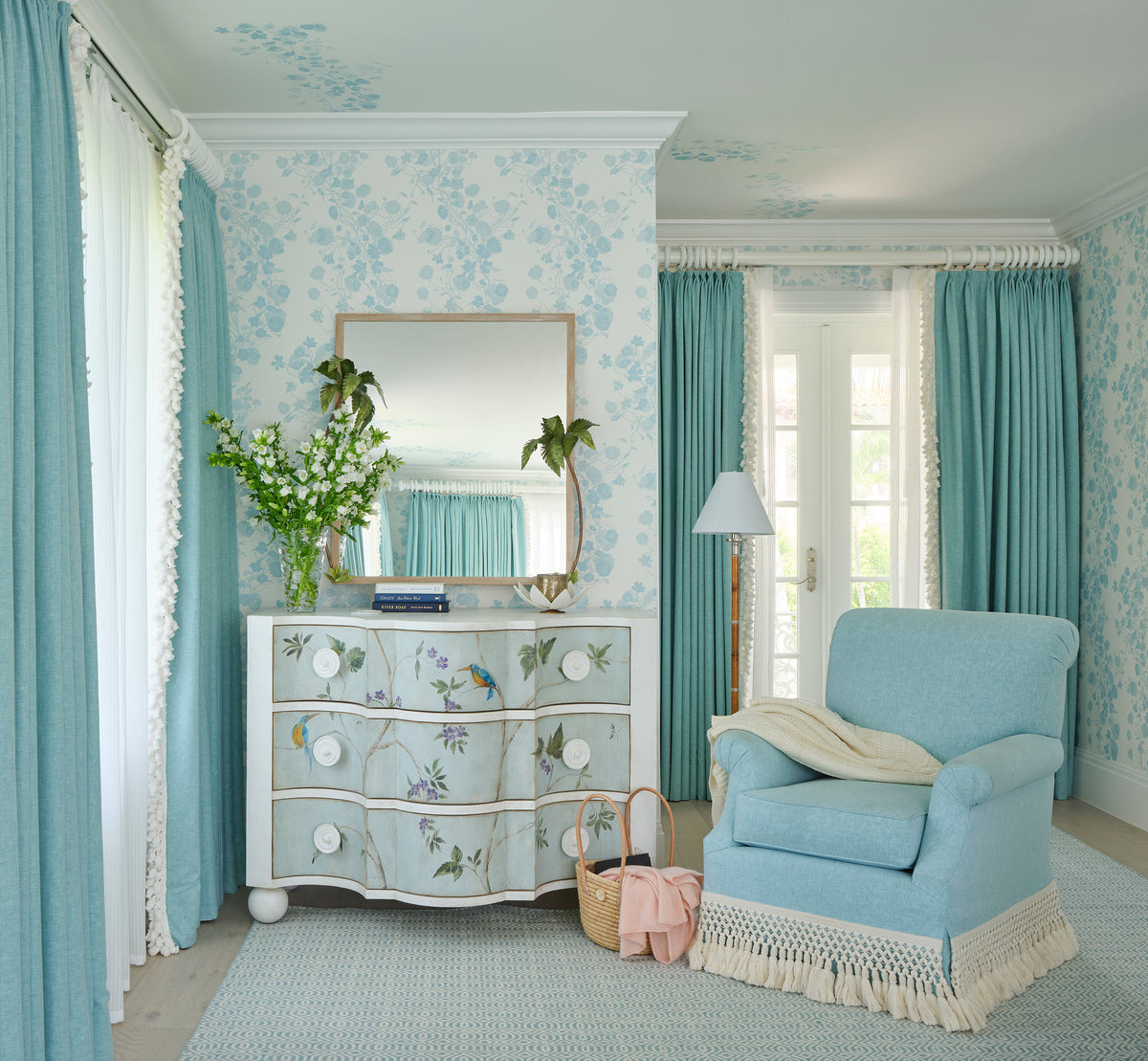 Bedroom corner with blue floral wallpaper and turquoise curtains, a hand painted serpentine chest under a gilt mirror, and a pale blue armchair with a fringed skirt
