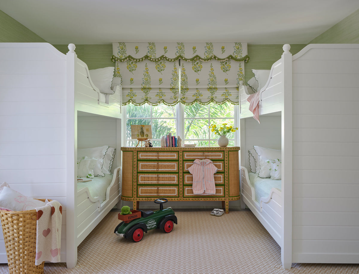 Children's room with two white four poster beds under a scalloped green valance, a caned chest between them and a toy car on the rug