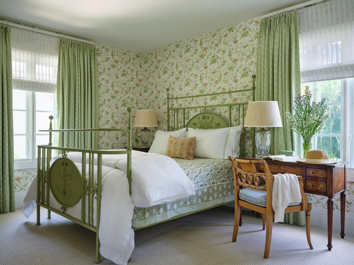 Bedroom in green floral wallpaper with matching curtains, a green painted iron bed and an antique writing desk with a straw hat on the chair