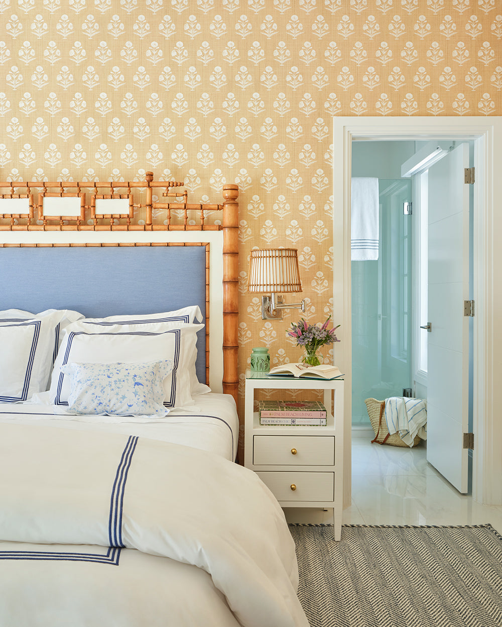 Bedroom with apricot patterned wallpaper and a faux bamboo headboard over navy trimmed white bedding, the bathroom beyond