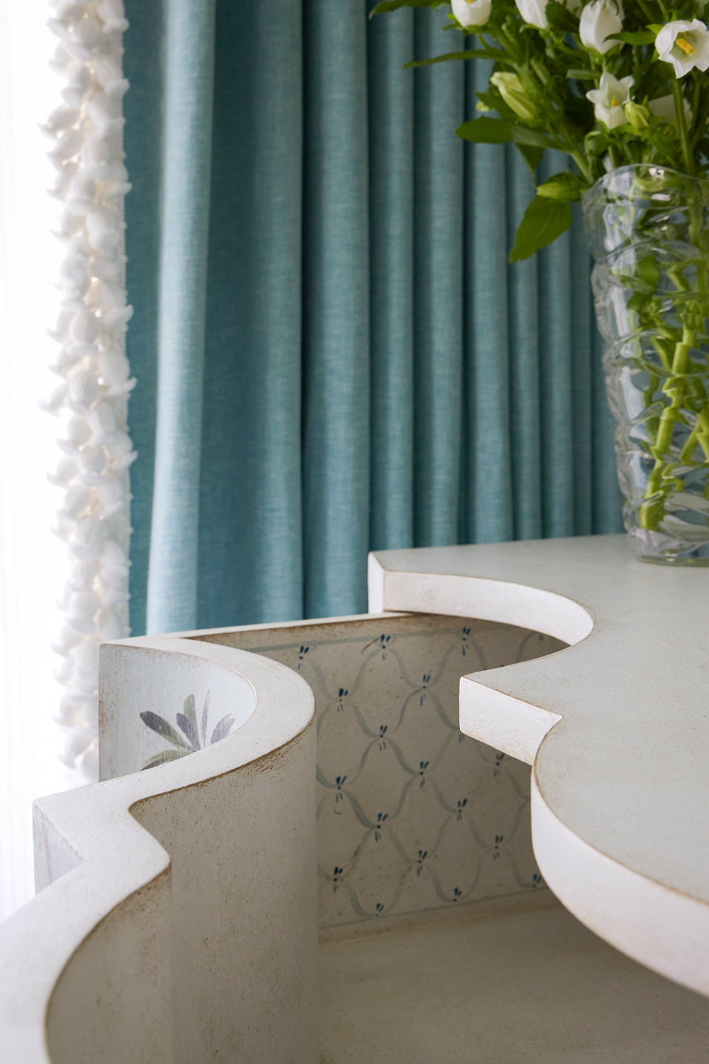 Curved plaster vanity with a trellis painted front against teal curtains, a tasselled mirror edge and white lilies in a glass vase