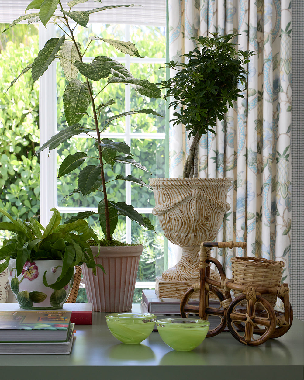 Window sill still life of a carved stone urn holding a topiary, potted ferns, a rattan bicycle ornament and green glass bowls