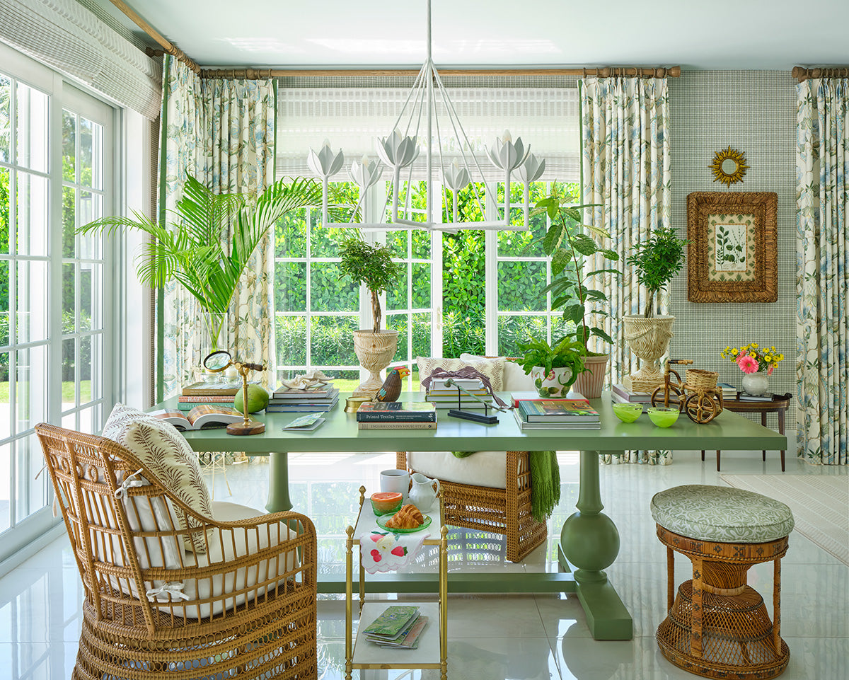 Garden room with a wall of windows, green and white floral curtains and a lotus pendant, a glass table laid with books and plants, rattan chairs and caned stools
