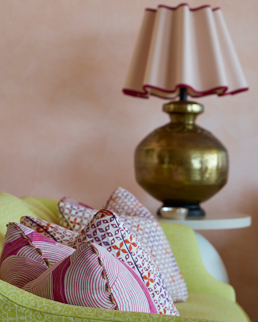 Detail of a brass lamp with a scalloped red trimmed shade over patterned pink cushions