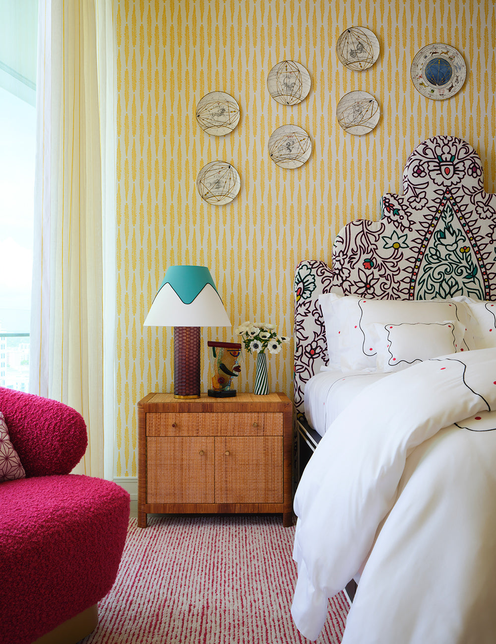 Bedroom with yellow patterned wallpaper, a scrolled embroidered headboard, a timber chest and a pink boucle chair