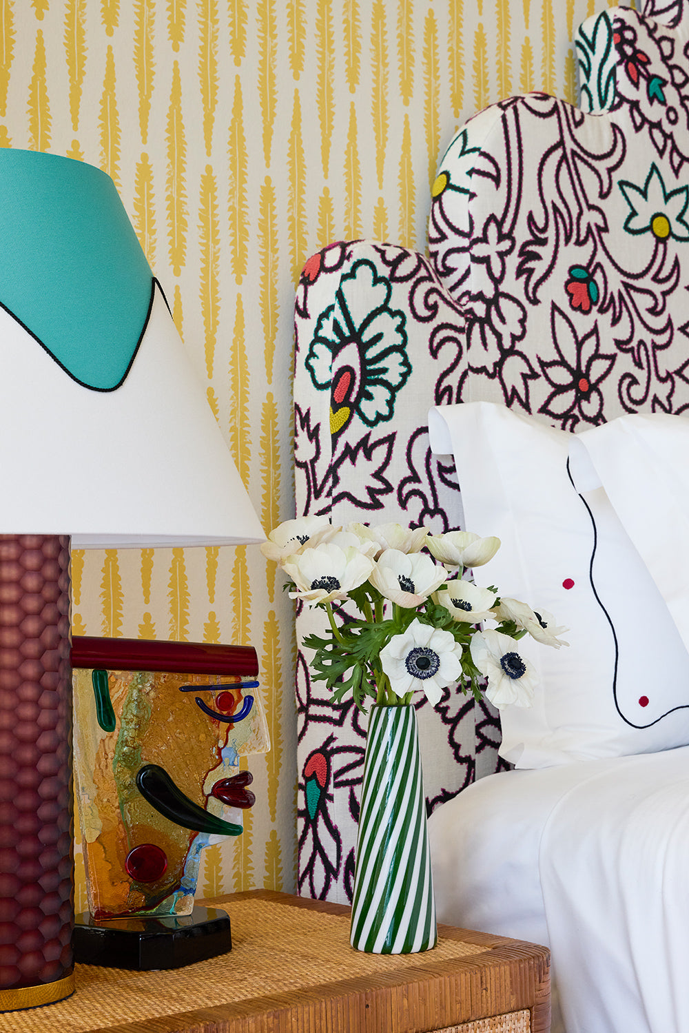 Headboard detail, embroidered scrolls against yellow wallpaper with a painted lamp and anemones in a striped vase