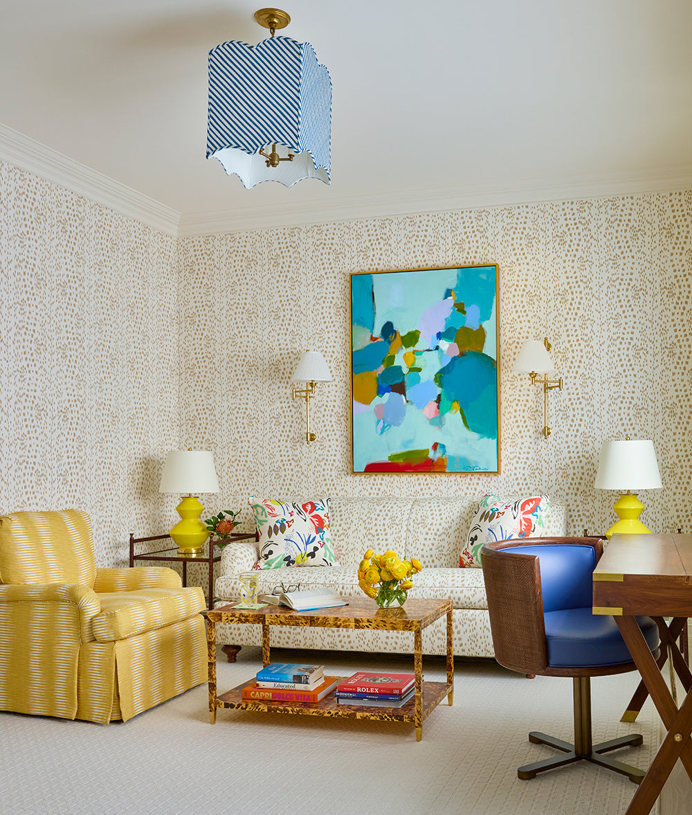 Sitting room with an abstract canvas between sconces, a floral sofa, a yellow skirted armchair and a blue desk chair