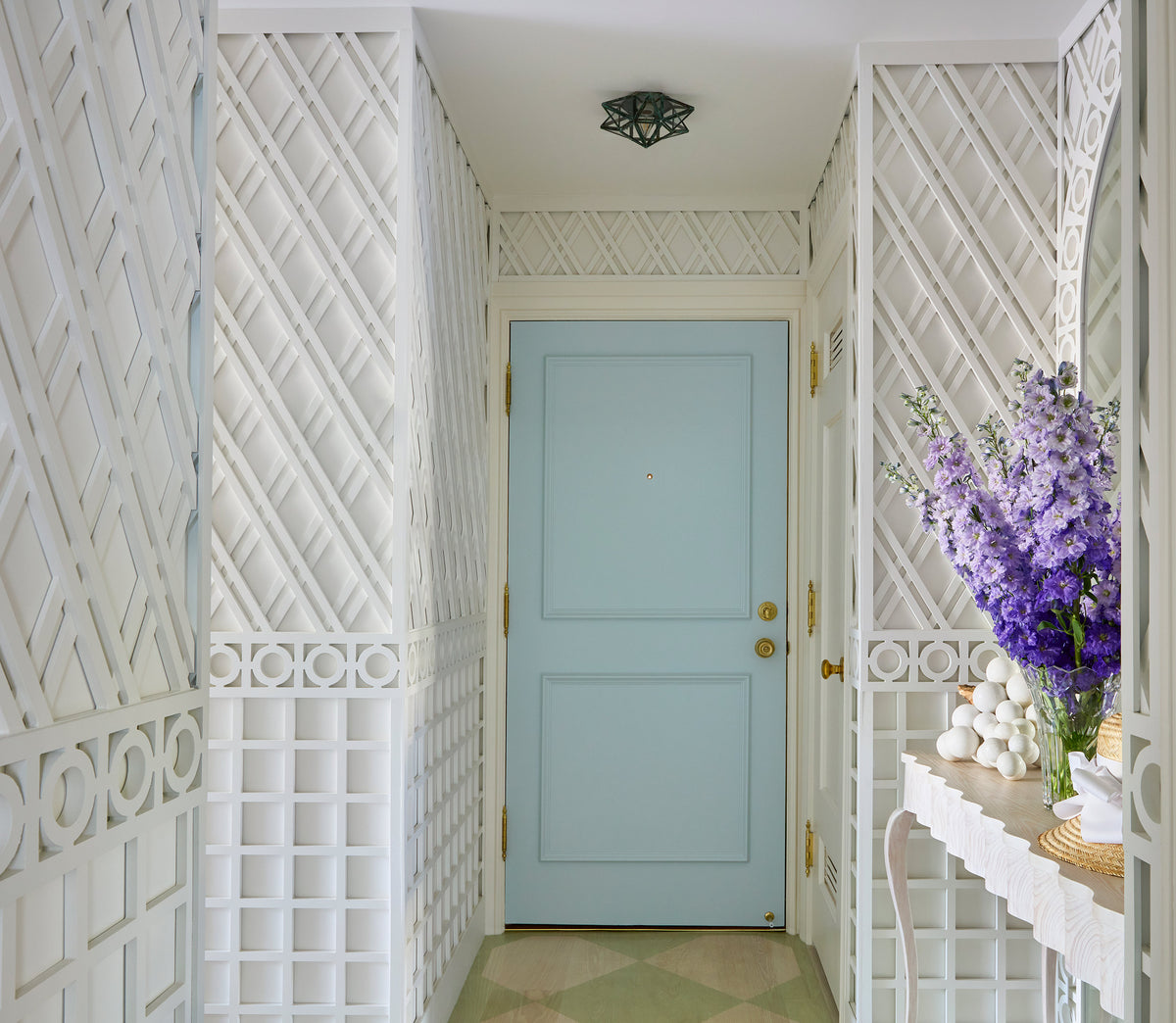 White trellis panelled hall with a pale blue door and delphiniums on a console
