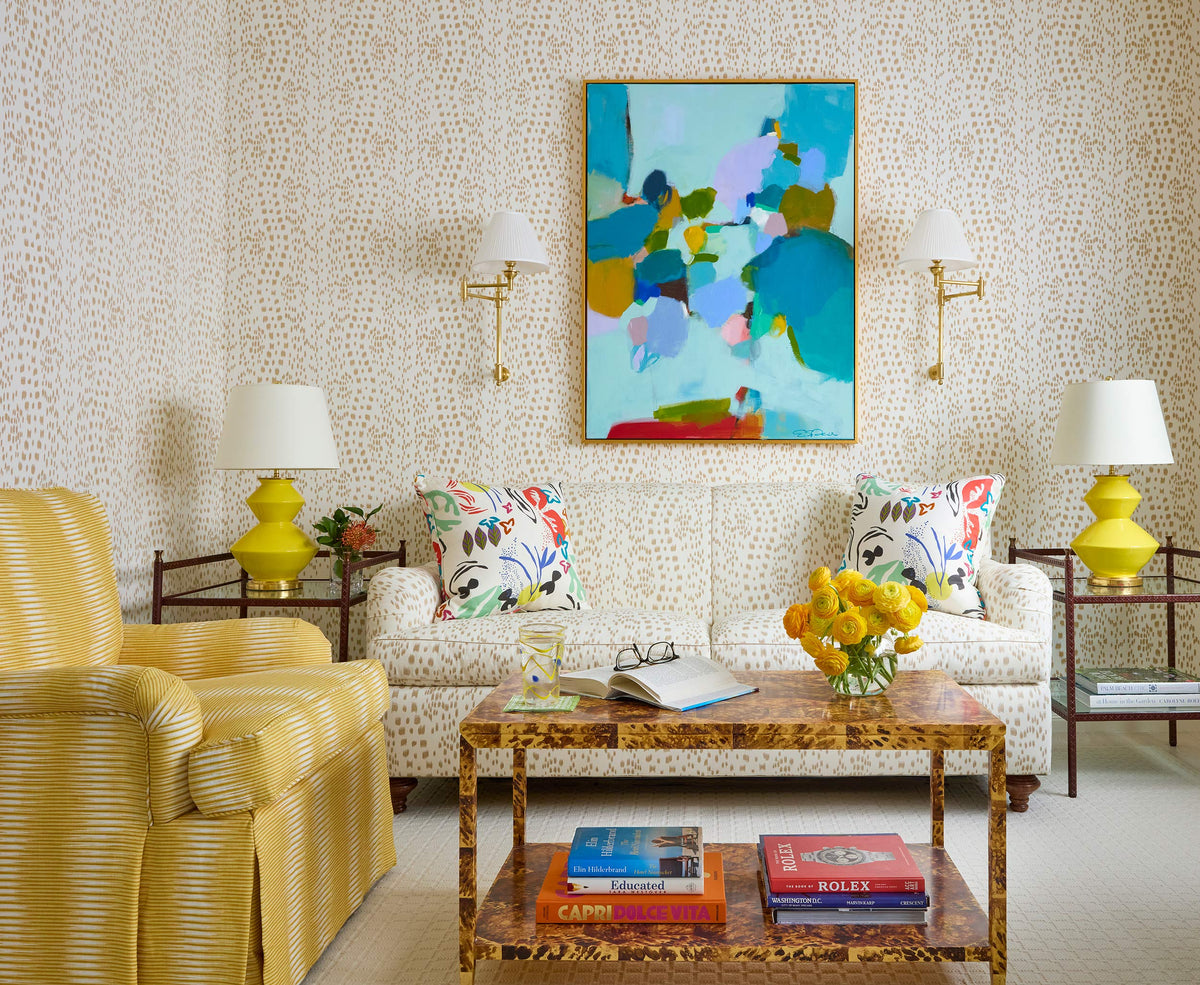 Sitting room with an abstract canvas between brass sconces, a floral sofa, yellow lamps and a tortoiseshell coffee table